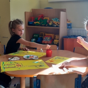 Troje dzieci siedzi przy stoliku i za pomocą kropek maluje obraz z żyrafą na pierwszym planie