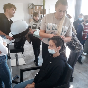 Zdjęcie przedstawia uczennicę siedzącą na fotelu podczas wykonywania fryzury przez fryzjera
